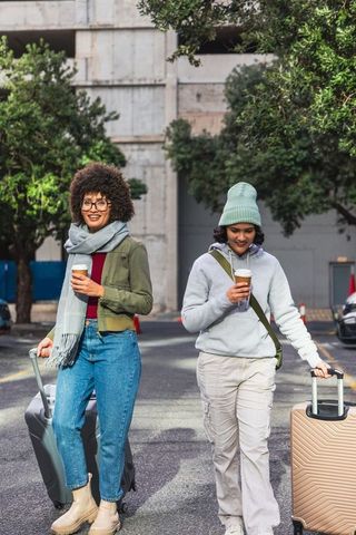 Diverse friends walking with hard-shell suitcases and coffee on urban street