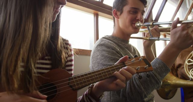 Two teenagers are engaged in playing musical instruments in a bright, naturally lit room. One is playing a ukulele, while the other is playing a trombone. Both have a look of concentration, emphasizing their interest and enjoyment in making music. It is a perfect representation of young musicians practicing, suitable for themes related to music education, youth engagement in arts, or indoor hobbies. Great visual for articles, educational materials, blog posts, or advertisements that promote musical learning classes or showcasing musical talent among teenagers.