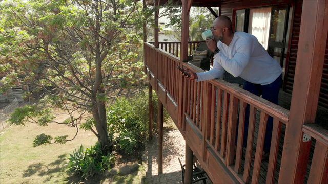 African American man enjoying tea while browsing smartphone on veranda of a wooden log cabin. Idea for concepts related to rural lifestyle, technology in nature, morning relaxation, peaceful getaways, remote work, and modern living in the countryside. Suitable for promoting vacation rentals, outdoor leisure activities, or technology usage in rural settings.