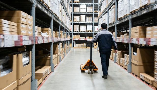 Warehouse worker pushing pallet jack down high-rack aisle stacked with cardboard boxes