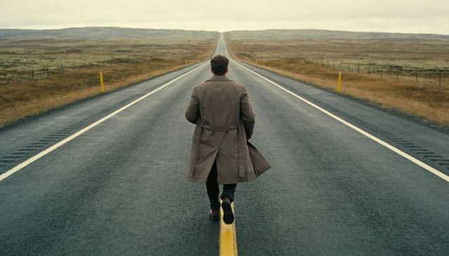Solitary man walking along endless wet road wearing trench coat toward distant horizon