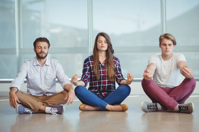 Three office workers sitting cross-legged on floor, practicing meditation together. Ideal for illustrating corporate wellness programs, stress relief techniques, and team-building activities in a professional environment.