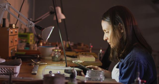 Female artisan concentrating while working on jewelry making at her workshop desk. Woman uses various tools and equipment in a cozy, well-organized workspace. Useful for promoting artisan craftsmanship, handmade product advertisements, or hobbyist features.