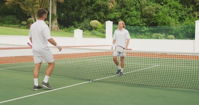 Two men, dressed in white sportswear, are playing tennis on an outdoor court surrounded by greenery. They are holding their racquets and appear to be engaging in a casual match or friendly competition. This could be used for content related to sports, fitness, exercise, recreational activities, summer fun, or promoting a healthy lifestyle.