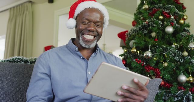 Senior man sits on couch in living room adorned with Christmas decorations, wearing Santa hat and using a digital tablet. Perfect for themes of holiday joy, virtual celebrations, and Christmas spirits. Ideal for promotions, articles, and advertisements focusing on festive technology use and staying connected during the holidays.