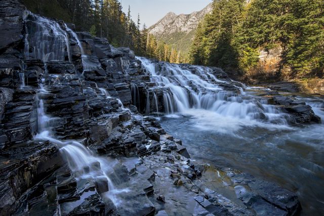 Water cascading down multiple rocky tiers in the Rocky Mountains, surrounded by lush forest and distant peaks. Perfect for nature, travel, and outdoor adventure themes, and highlighting serene and pristine natural landscapes.