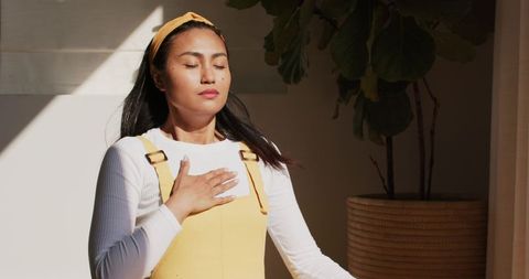 Asian woman practicing mindful breathing in sunlit room wearing yellow pinafore
