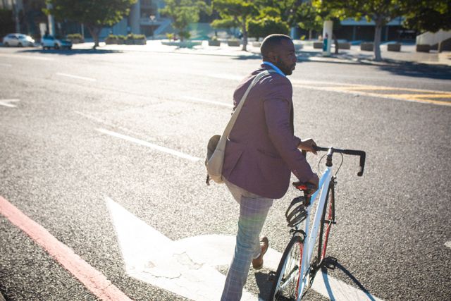 African American businessman crossing street with bicycle in morning light. Ideal for content related to urban commuting, eco-friendly transportation, professional lifestyle, and city living. Can be used in articles, blogs, and advertisements promoting sustainable travel, business attire, and urban life.