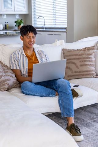 Young asian man working on laptop from sofa in bright modern open-plan apartment