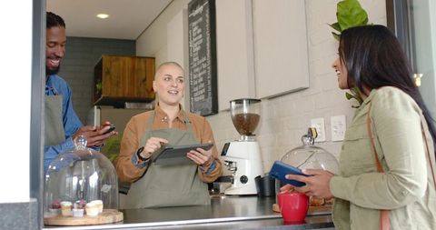 Baristas serving indian customer at cafe counter holding tablet offering espresso, cupcakes