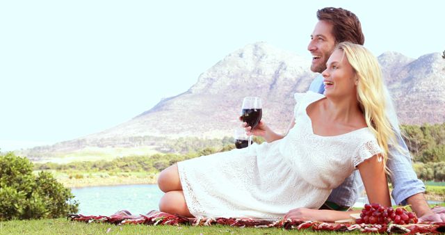 A young Caucasian couple is enjoying a romantic picnic with a scenic mountain backdrop, with copy space. They are sharing a moment of happiness with glasses of wine, embodying leisure and connection with nature.