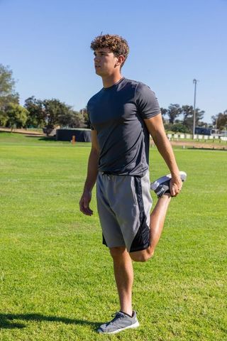 Young male athlete performing standing quadriceps stretch on grassy field wearing running shoes