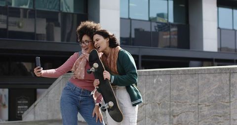 Two friends taking selfie with skateboard on urban plaza laughing casual street style