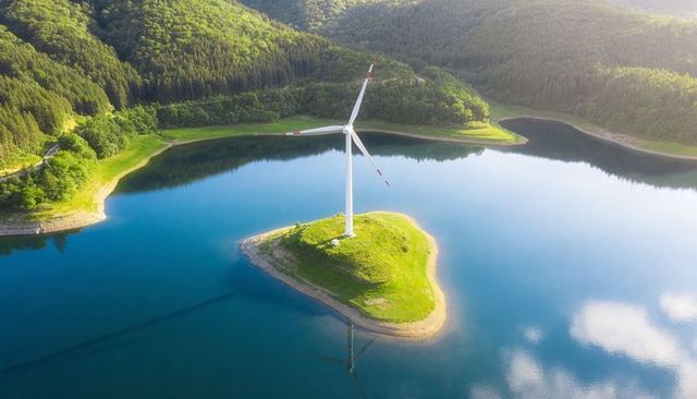 Towering wind turbine rising from grassy islet in mirror-blue reservoir, forested valley