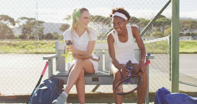 Image of happy diverse female tennis players sitting, talking and resting after match. professional tennis training, sport and competition concept.