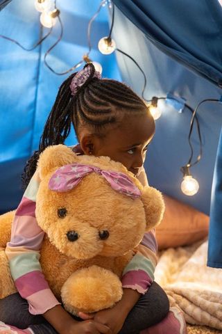 African american girl hugging teddy bear in cozy blue fort with warm string lights