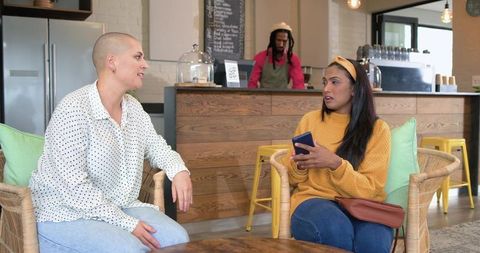 Multicultural women chatting in cozy cafe sitting in wicker chairs with smartphone