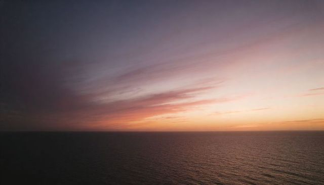 Glowing pastel sunset over open ocean with wispy clouds and tranquil reflective horizon at dusk