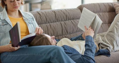 Two women reading together on cozy sofa, wearing denim jackets and relaxing at home