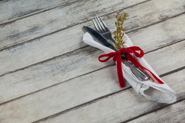 Festive cutlery wrapped in a white napkin with a red ribbon and gold ornament placed on a rustic wooden table. Ideal for holiday dining themes, restaurant promotions, festive table settings, and celebration invitations.