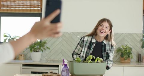 Young woman smiling while tending indoor planter in sunlit modern kitchen, casual plaid