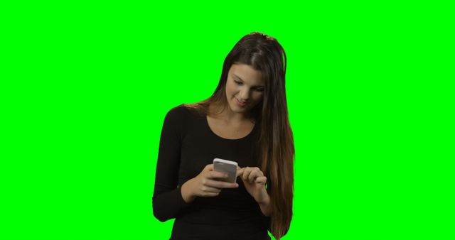 Young woman standing and using a smartphone with a green screen background. Ideal for projects that require isolation of the subject for chroma key compositing. Usage includes tech articles, website backgrounds, apps, digital advertising, social media promotions, or tutorials about mobile device usage.
