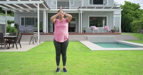 Mature woman practicing yoga on backyard lawn by pool deck for home fitness and mindfulness