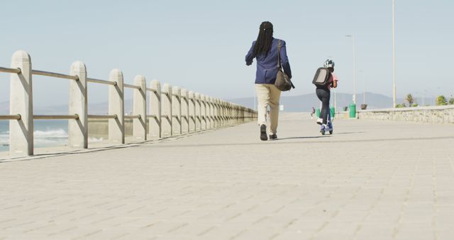 Father walking with child riding scooter along seaside promenade on a sunny day. Ideal for concepts related to family time, outdoor activities, father's day, urban lifestyle, and exercise. Suitable for advertisements or articles about parenting, health, travel, and leisure.