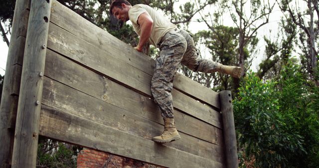 Army soldier actively climbing over a large wooden obstacle wall during an outdoor military training session. Wearing camouflage pants and combat boots, displaying physical fitness and determination. Useful for showcasing military activities, physical challenges, teamwork in armed forces, and themes of strength and endurance.