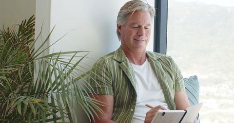 Senior man journaling by sunlit window in green striped shirt, relaxed reflective lifestyle