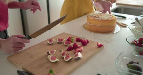 Friends spreading frosting while slicing figs and strawberries on wooden cutting board