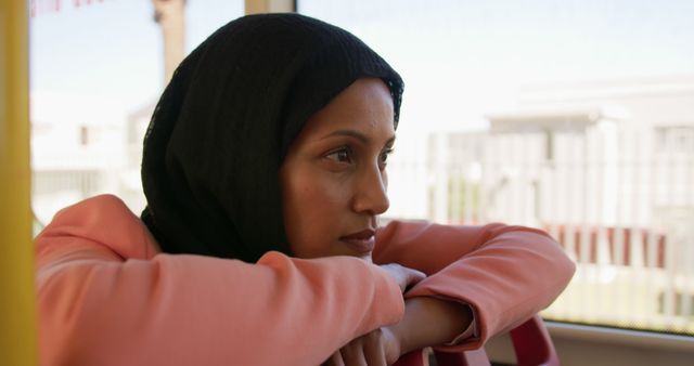 Muslim woman in a hijab appears thoughtful while looking out a window on public transportation. Ideal for themes of introspection, travel, diversity, public transport, and personal contemplation.