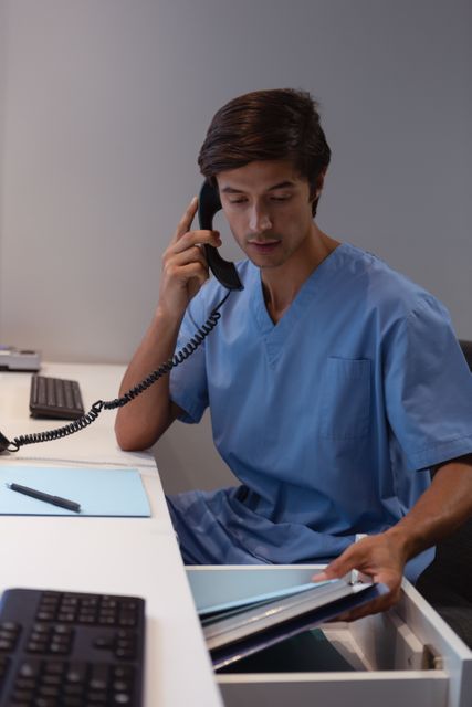 Caucasian male surgeon in blue scrubs working at his desk in a hospital, talking on the phone and looking through documents in a drawer. Ideal for use in healthcare, medical, and professional contexts, illustrating a busy medical professional multitasking in an office environment.