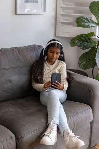 Indian girl relaxing on gray couch using smartphone and headphones, indoor plant, natural light