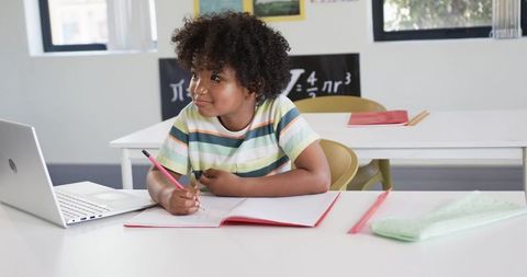 Elementary Student Writing in Notebook While Using Laptop for Remote Learning in Classroom