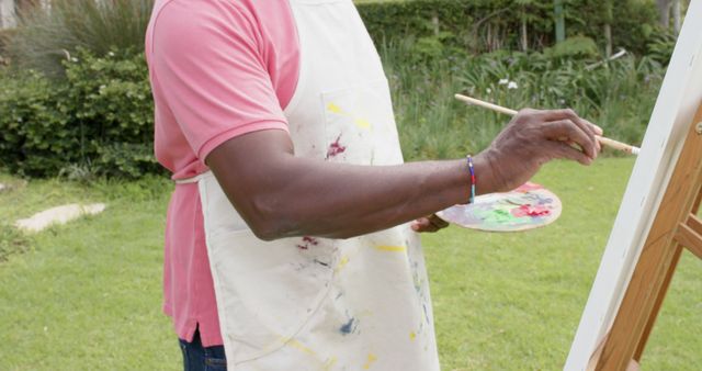 Man painting on easel outdoors in garden, wearing apron and holding palette and paintbrush. Ideal for articles on artistic hobbies, creativity, outdoor activities, and relaxing pastimes.