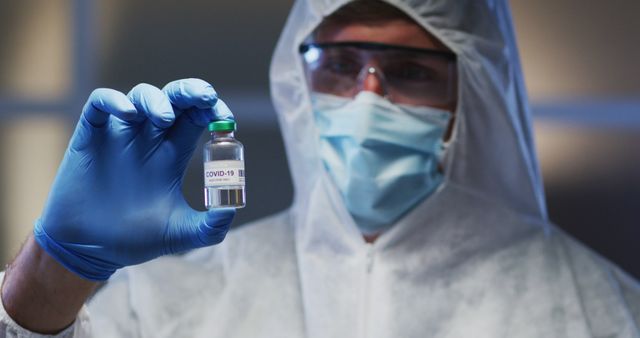 Scientist holding COVID-19 vaccine vial while wearing protective gear. Ideal for illustrating vaccine research, pandemic response efforts, coronavirus updates, and scientific breakthroughs. Useful for health campaigns, educational materials, and news articles related to COVID-19 vaccinations.