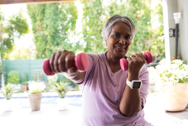 Senior woman exercising with pink dumbbells in backyard, promoting active and healthy lifestyle. Ideal for use in fitness, wellness, and health-related content, showcasing strength training and outdoor workouts for elderly individuals.