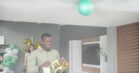 Man unfolding gold birthday banner in modern living room with teal balloons 30th