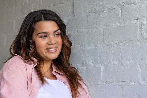 Smiling young woman leaning on gray brick wall, wearing pink shirt and white ribbed top
