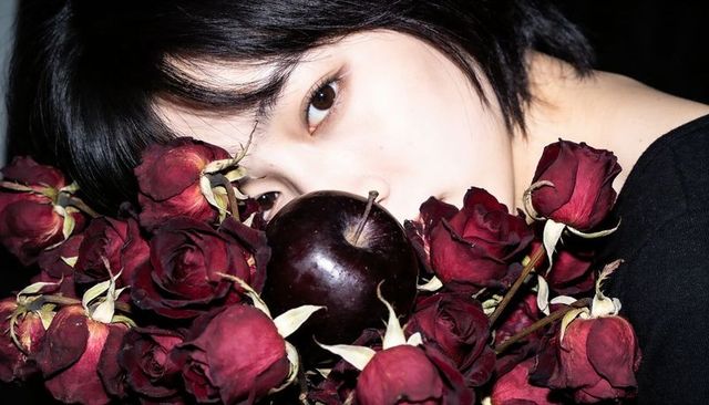 Moody closeup portrait peeking behind wilted crimson roses with glossy purple apple