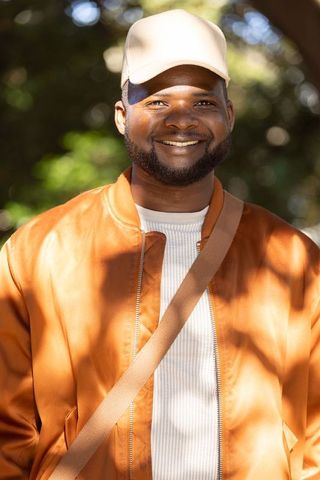 Smiling man wearing beige cap and orange bomber jacket on sunlit park path