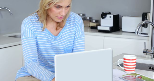 Blonde woman is working on a laptop in a modern kitchen with a striped coffee mug beside her. Ideal for illustrating remote working, home office setups, freelance work, telecommuting, and productivity in home environments.