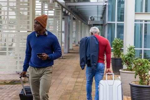 Two african american men walking through modern glass corridor pulling rolling suitcases