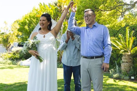 Mature couple celebrating garden wedding holding bouquet and raising hands in sunshine