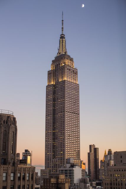 Empire State Building illuminated under dusk sky with visible moon. Ideal for travel brochures, posters, architectural magazines, and websites showcasing New York City's landmarks.
