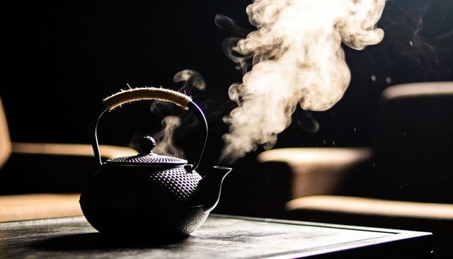 Steaming cast-iron teapot releasing tall vapor plume on dark table with wrapped handle
