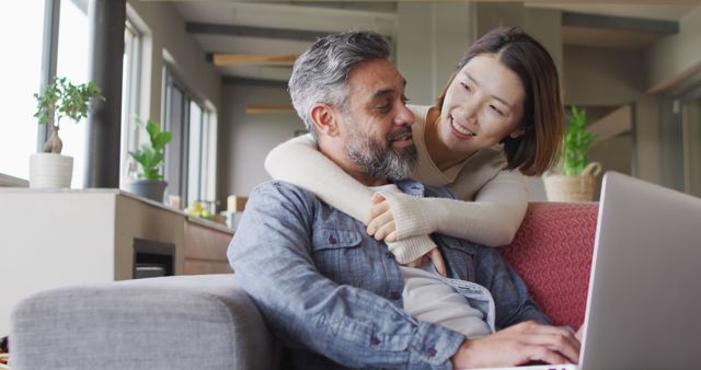 Asian woman and mixed-race husband sharing intimate moment in living room while using laptop. Ideal for promoting home lifestyle, family technology integration, and relationship content.