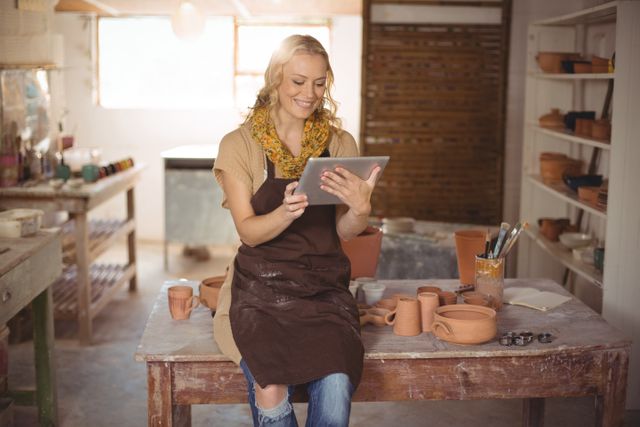 This image depicts a female potter sitting on a table using a digital tablet in her ceramic workshop. She is surrounded by handmade pottery pieces, paints, and brushes, indicating an active and creative environment. The potter's smile suggests she is content and engaged in her craft. This image is ideal for illustrating concepts related to technology in art, small businesses, creative workshops, and the blending of traditional and modern techniques.