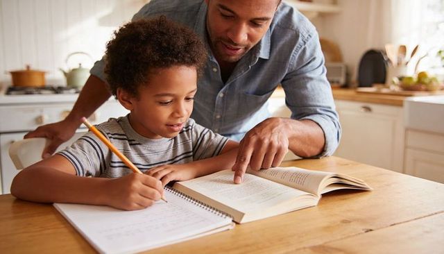 Father guiding child doing homework at sunlit kitchen table reading and writing together